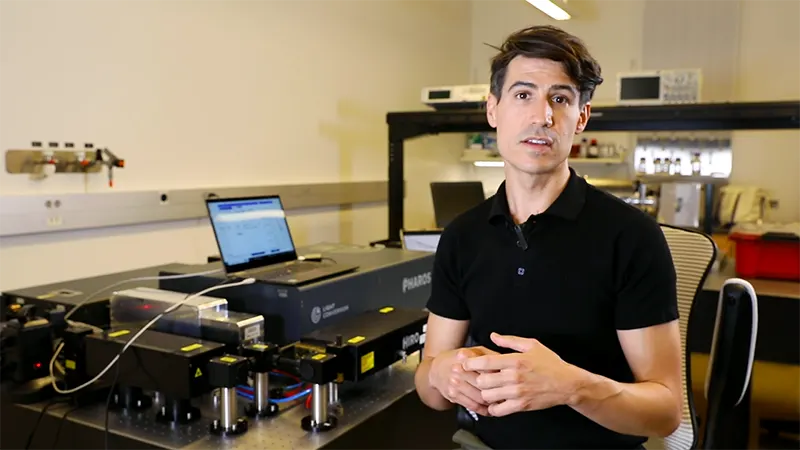 A researcher speaking to camera in front of laser optics lab equipment
